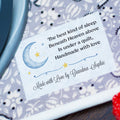 A close-up of a personalized quilt with a printed nursery poem on a white label, being ironed on a patterned fabric surface with a teal iron visible on the left and a red textured fabric on the right.