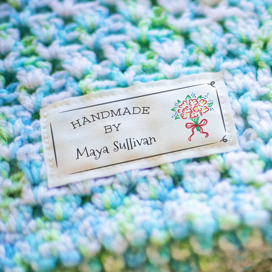 A close-up of a handmade crocheted blanket in variegated light blue, mint green, and cream yarn, featuring a cream fabric label sewn onto the side that reads 'HANDMADE BY Maya Sullivan' with a small floral illustration.