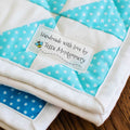 A folded quilted fabric item with alternating white and light blue polka-dot diamond panels, shown on a wooden surface. A sewn label reads, 'handmade with love by Tessa Montgomery with a bumble bee image