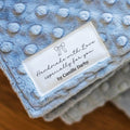 A close-up of a light blue textured baby blanket with a white fabric label that reads 'Handmade with Love especially for you by Camille Darby' on a wooden surface.
