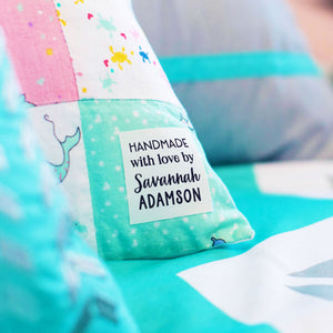 A close-up of a handmade patchwork pillow with a white fabric label that reads 'HANDMADE with love by Savannah ADAMSON'. The pillow features various colorful fabric panels, including pink, mint green, and white, and is styled on a bed with a turquoise blanket in the foreground.